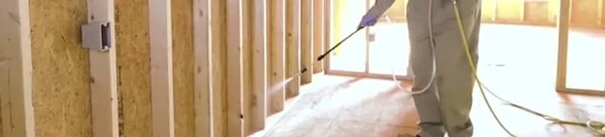 Termite treatment technician applying spray along framing and unfinished walls in Staten Island, NY