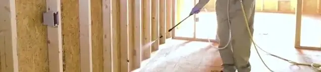 Termite treatment technician applying spray along framing and unfinished walls in Staten Island, NY